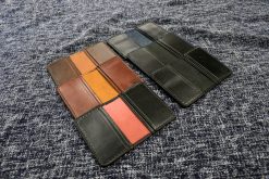 Three different colors of Grommet's Leathercraft Bernie Bi-Fold Black Leather Wallets sitting on top of a blue cloth.